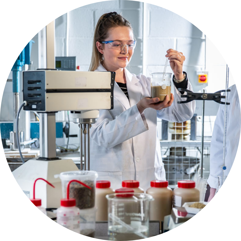 Women in white lad coat, wearing protective eye wear, holding a beaker and string contents with glass tube.  Standing in a lab, surrounded by scientific equipment and materials