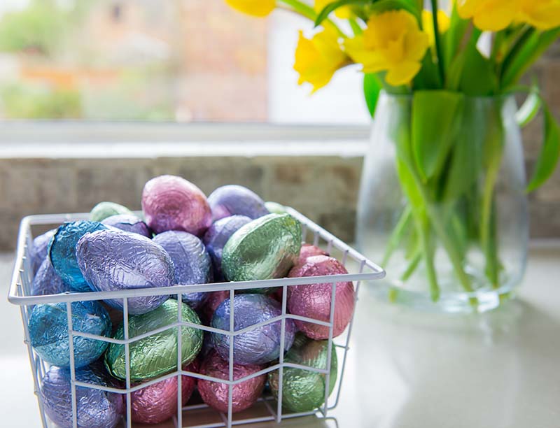 Chocolate Easter eggs in a wire basket with a vase of daffodils behind sign post.jpeg