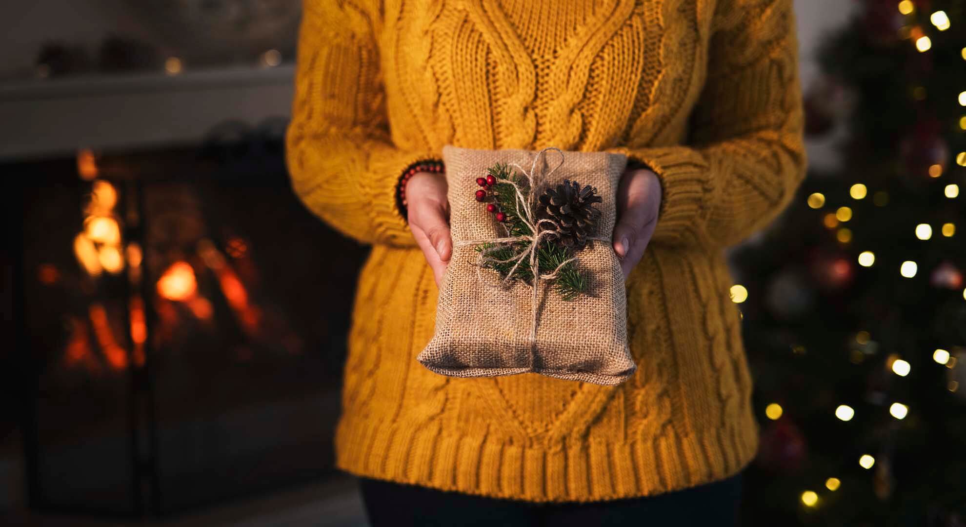 Person standing beside a Christmas tree holding a gift wrapped in hessian fabric and twine