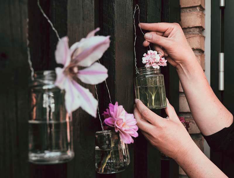 Person hanging glass jam jars with flowers in them onto a fence