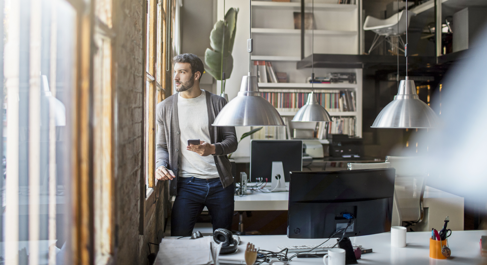 A person standing in an office looking out a window, holding a mobile phone